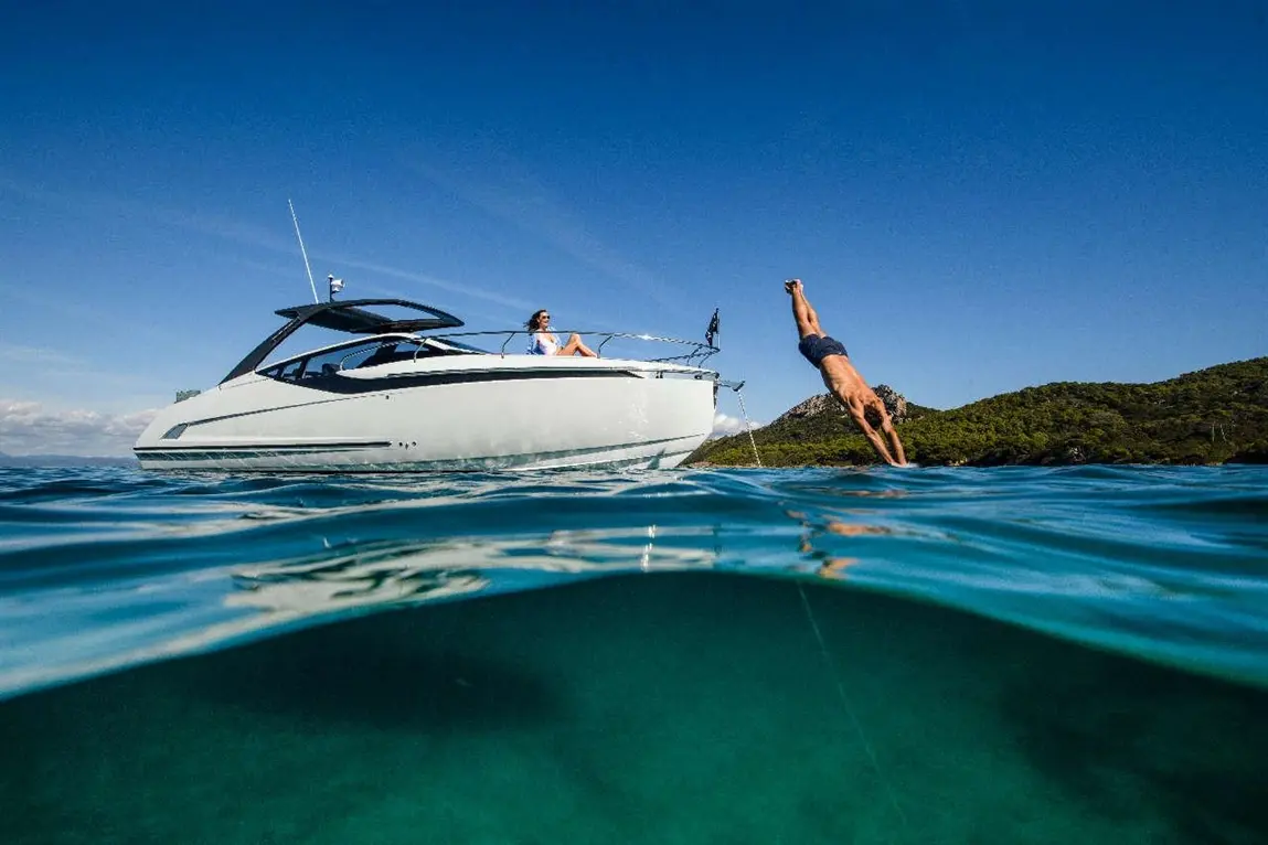 A man dives into clear water near a sleek boat where a woman relaxes, surrounded by a scenic landscape and blue sky.